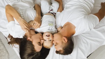 Pareja con camisetas blancas tumbados en la cama y besando a su bebe entre ellos